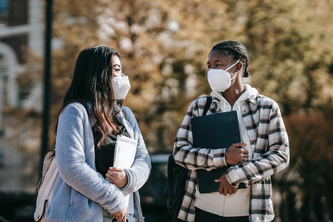 Two female young adults stand talking, wearing N95 masks and holding books to their chests.